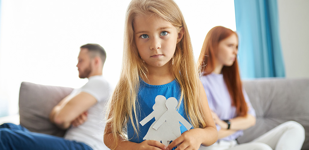 Meisje kijkt in camer met ouders boos achter zich wegkijkend van elkaar op de bank. | Hoe kun je steun bieden aan gescheiden ouders en kinderen?