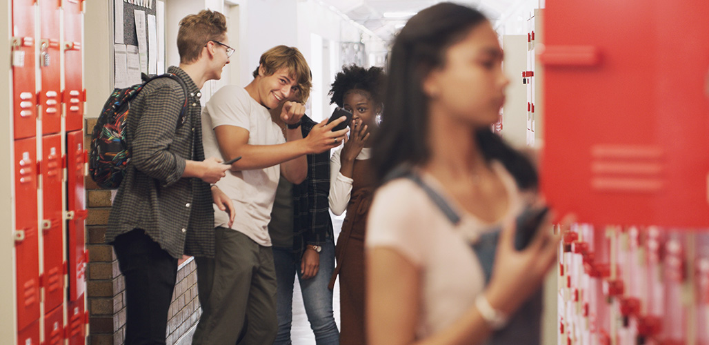 Pubers kijken op mobiel en wijzen naar meisje die bij haar eigen kluisje staat. | Actueelbericht: Online veiligheid en kansengelijkheid onder grote druk 