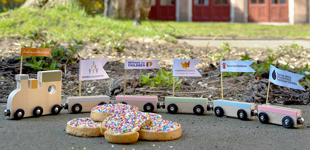 Foto van beschuit met muisjes en een houten trein op achtergrond met daarop alle deelnemers aan het Kinderrechtencollectie dat doorgaat als stichting.