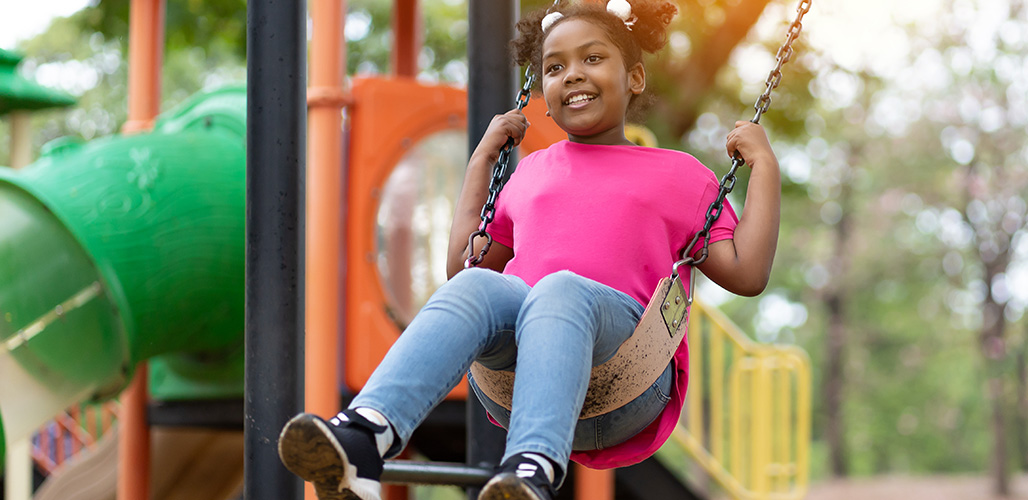 Meisje op schommel met op achtergrond een speeltoestel. - Actueelbericht: Wat kan ondersteunend personeel in de maatschappelijke opvang doen voor kinderen?
