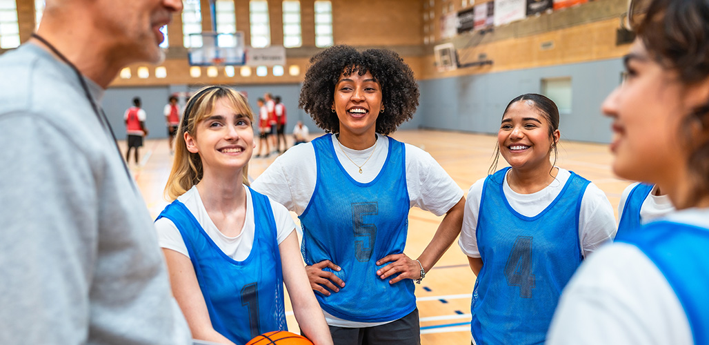 Sportieve groep jonge meiden die bij de coach voor uitleg staan voor hun basketbalwedstrijd | Actueelbericht: Sportfonds voor Mentale Gezondheid helpt jongeren