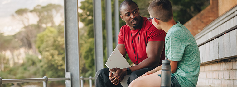 Mentaal welzijn en jongeren | Buurtcoach in gesprek met jongen bij sportveld op tribune