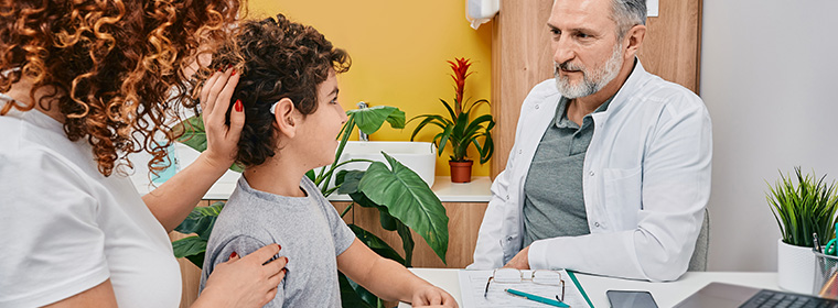 Arts luistert naar jongen die op consult komt. Moeder steunt de jongen op achtergrond.